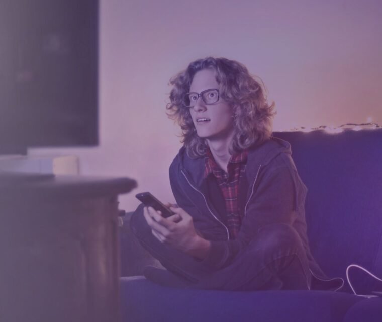 Young man in a living room using a smartphone and watching TV, enjoying seamless streaming with the best IPTV service and premium channels.